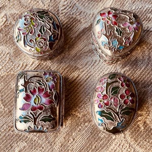 May include: Four silver-toned trinket boxes with floral enamel designs. The boxes are in various shapes: round, heart-shaped, rectangular, and oval. The enamel colors include pink, green, blue, and yellow.