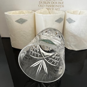 May include: A set of clear crystal old fashioned glasses featuring a geometric cut pattern. The packaging box is labeled 24% LEAD CRYSTAL DUBLIN DOUBLE OLD FASHIONEDS 4 PIECE SET. Three glasses are wrapped in white tissue paper.