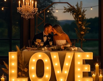 May include: A romantic wedding scene with a large illuminated "LOVE" sign in the foreground. A couple is seated at a table, with candles and floral arrangements. A chandelier and string lights add to the ambiance.