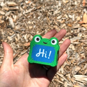 May include: A green frog-shaped badge with a blue rectangular center featuring the white text Hi!. The frog has two prominent eyes with black pupils and a small black smile. The design has a textured finish and a bright, vibrant green border.
