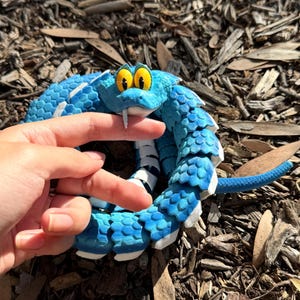 May include: A blue and white toy snake with a cartoonish face, featuring large yellow eyes and a white tongue. The snake is coiled and has a segmented body. The toy is resting on a bed of wood chips.
