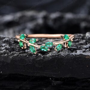 May include: A rose gold ring with a leaf design, set with emerald green marquise-cut gemstones. The ring has a slender band, and the leaves are arranged in a delicate pattern. The ring is displayed against a dark, textured background.