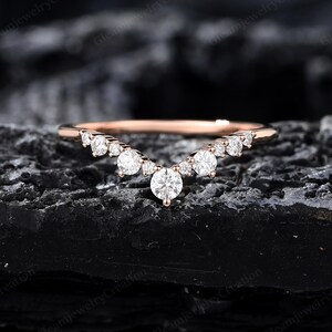 May include: A rose gold ring with a curved design, set with multiple round, clear gemstones. The ring features a central, larger stone, with smaller stones arranged in a graceful arc. The ring is displayed on a dark, textured surface.