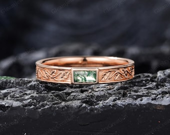 Puede incluir: Un anillo de oro rosa con una gema verde rectangular. La banda presenta un diseño floral grabado. El anillo está sobre una superficie oscura y texturizada.
