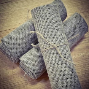 May include: Several rolled gray burlap fabric napkins tied with twine, arranged on a wooden surface. The napkins are tightly rolled and secured, showcasing the rustic texture of the burlap material. The neutral color palette suggests a natural, eco-friendly theme.
