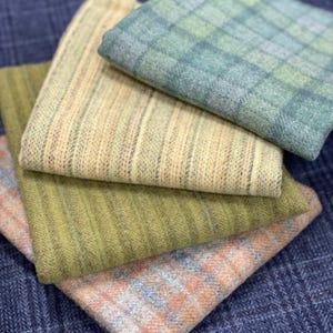 May include: A collection of folded fabric swatches in various colors and patterns. The swatches include a green plaid, a peach and blue plaid, a ribbed beige, and a ribbed green fabric. The background is a blue fabric with a subtle pattern.