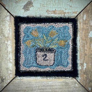 May include: A square, hand-tufted rug with a floral design. The rug features a light blue background with yellow flowers in a white pot, and the number "2". The rug is framed by a black border.