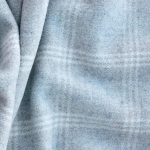 May include: A soft, light blue fabric featuring a subtle white plaid pattern. The material has a fuzzy, brushed texture with visible folds and drapes that create varying shades of blue across the surface of the textile.