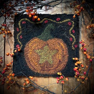 May include: A square, handmade rug with a pumpkin design. The pumpkin is orange and brown with a green stem and leaf. The background is black, and the rug is framed with a decorative border and red accents. The rug is a fall-themed decorative item.