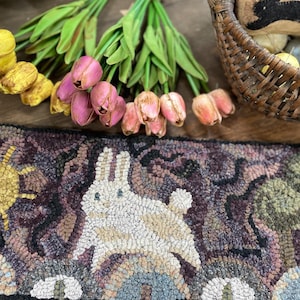 May include: A hand-hooked rug with a rabbit design, featuring a sun and trees, in shades of purple, brown, and cream. The rug is decorated with a bunny, sun, and trees. Tulips and a basket with Easter eggs are in the background.