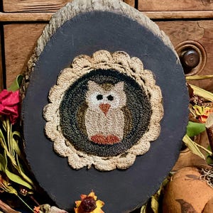 May include: Oval, dark gray wooden plaque with a stitched owl design. The owl is centered within a circular, beige, crocheted border. The owl has white eyes, a brown body, and orange feet. The plaque has a natural wood edge.