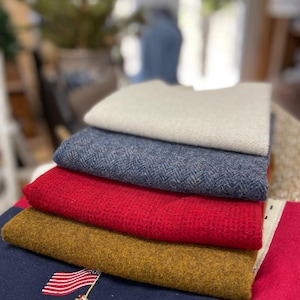 May include: A stack of colorful fabric swatches. The top swatch is a light beige, followed by a blue herringbone pattern, a vibrant red, and a mustard yellow. The bottom swatch is navy blue with an embroidered red truck and American flag.