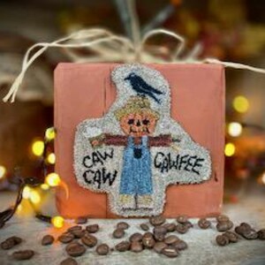 May include: A square, orange-colored decorative item with a stitched design of a scarecrow and a crow. The scarecrow has a pumpkin head and is wearing blue overalls. The text "CAW CAW CAWFEE" is stitched below the scarecrow. Coffee beans are scattered in front.