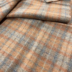 May include: A piece of woven fabric featuring a plaid pattern in shades of tan, light brown, and muted blue. The material has a soft, slightly fuzzy texture with visible fibers throughout, displaying a classic grid design across the surface.