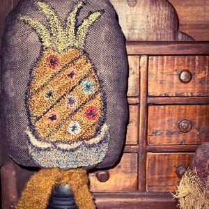 Puede incluir: Un cojín decorativo con forma de piña sobre un fondo gris oscuro. La piña está bordada con hilos naranjas, amarillos, azules y rojos. Se apoya sobre un soporte negro con una base marrón texturizada. Un mueble de madera está en el fondo.