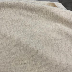 May include: A piece of off-white fabric with a visible woven texture and a subtle heathered appearance. The material features a soft, slightly fuzzy surface with small, darker flecks distributed throughout the light beige base color.