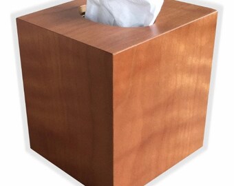 May include: A wooden tissue box cover in a light brown color with a square shape. The tissue box cover has a slot for tissues to be pulled out.