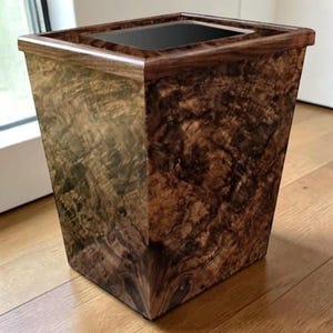 May include: A wooden wastebasket with a dark, patterned finish. The rectangular container has a dark interior and a contrasting, darker wood trim around the top edge. The wastebasket is set on a wooden floor.