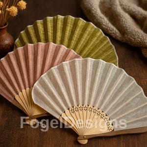 Puede incluir: Tres abanicos de mano en tonos rosa, verde y beige, con mangos de madera. Los abanicos están desplegados, mostrando su diseño de tela plisada. La marca "Fogel Designs" está impresa en la parte inferior de los abanicos.