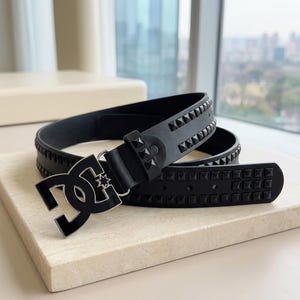 Peut inclure: Une ceinture noire dotée d'une grande boucle avec le logo DC stylisé et un contour argenté. La sangle est ornée de deux rangées parallèles de petits clous noirs en forme de pyramide carrée, incluant un clou assorti sur le passant de ceinture.