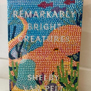 May include: A rectangular book cover art piece covered in small, iridescent rhinestones. The design features an underwater scene with a large orange octopus, coral, and fish. The text reads REMARKABLY BRIGHT CREATURES by SHELBY VAN PELT.