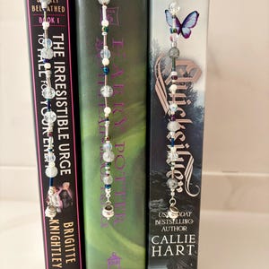 May include: Three beaded bookmarks with silver hooks are inserted into the pages of books. The bookmarks feature clear, white, and colored beads. The books are titled "The Irresistible Urge", and "Harry Potter", and "Quicksilver".