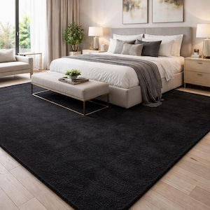 May include: A large, rectangular, solid black area rug with a plush, textured pile. The rug is positioned on a light-coloured wooden floor, covering a significant portion of the floor space in front of a bed and a bench in a bedroom setting.