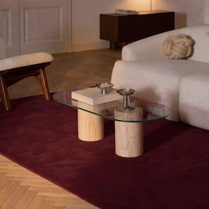 May include: An oval coffee table featuring a clear glass top supported by two cylindrical beige travertine stone bases. The table is positioned on a deep burgundy area rug, with two small silver metallic bowls placed on the glass surface.