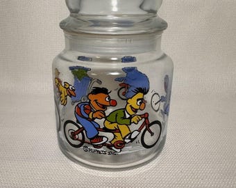 Könnte beinhalten: Ein durchsichtiger Glasbehälter mit passendem Glasdeckel, verziert mit einer bunten Grafik von Sesame Street-Figuren auf Fahrrädern. Der Aufdruck zeigt Bert und Ernie auf einem Tandem, Bibo und das Krümelmonster sowie den Text Muppets Inc.