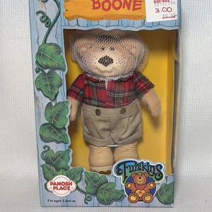May include: A Boone teddy bear plush toy inside a cardboard box. The bear wears a red and green plaid shirt, tan shorts, and tan shoes, with a white mesh net over its head. The box features green ivy illustrations and the text BOONE and Twiskins.
