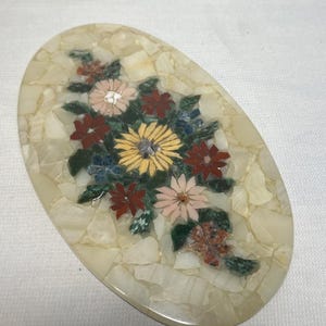 May include: An oval-shaped decorative plaque featuring a mosaic inlay of various flowers. The background consists of cream-colored stone fragments. The floral design includes yellow, pink, dark red, and blue stone pieces with green leaves.