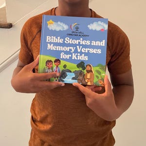 Op de afbeelding: Een persoon houdt een boek vast met de titel "Bible Stories and Memory Verses for Kids". De blauwe kaft toont cartoon illustraties van kinderen, een waterval en een bijbelse sc&egrave;ne. De titel van het boek staat in grote, blauwe letters.
