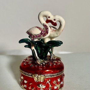 May include: A decorative red trinket box with a pair of white swans with pink jeweled accents. The swans are positioned above green leaves. The box is embellished with rhinestones and gold trim.