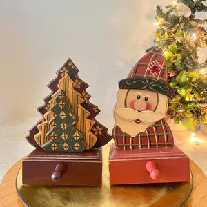 Könnte beinhalten: Zwei hölzerne Weihnachtsdekorationen mit Schubladen. Eine zeigt gestapelte, geschichtete Weihnachtsbäume in Dunkelrot, Gelb und Grün mit Sternmustern. Die andere ist eine Weihnachtsmannfigur mit rot karierter Mütze und Mantel.