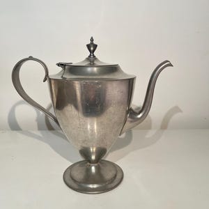 May include: A silver-toned metal teapot with a smooth, tapered body, a curved handle, and a long, slender spout. The teapot features a circular base and a removable lid topped with a small, pointed finial. The surface shows a matte finish.
