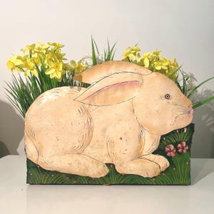 May include: A flat, beige rabbit-shaped decorative piece with painted details, resting on a green base with small red flowers. The rabbit has long ears and visible whiskers. Behind the rabbit, a cluster of bright yellow daffodils is visible.