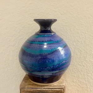 May include: A handcrafted ceramic vase with a round body and a dark blue neck. The vase features horizontal bands of blue and teal, creating a textured, layered effect. The base is a light brown color. This decorative vase is perfect for displaying flowers.