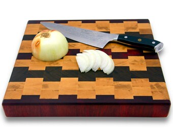 Op de afbeelding: Een vierkant houten snijplank met een schaakbordpatroon van licht en donker hout. De plank heeft een donkerrode rand. Een witte ui en een paar uisnijdtjes liggen op de plank, samen met een zilveren koksmes.