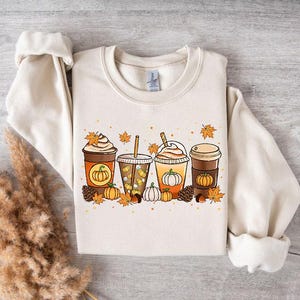 May include: Cream-colored sweatshirt featuring a fall-themed graphic. The design includes four different coffee drinks with pumpkins, leaves, and pinecones. The sweatshirt has a crew neck and long sleeves.