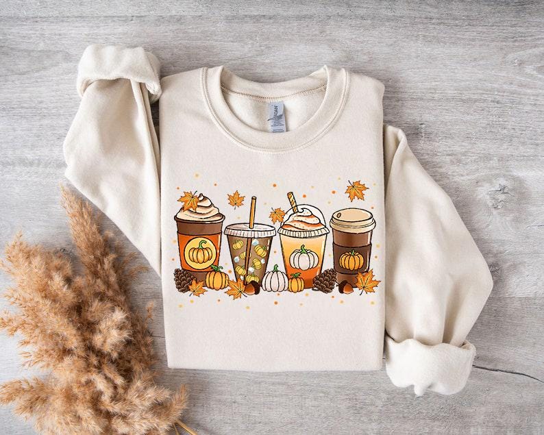 May include: Cream-colored sweatshirt featuring a fall-themed graphic. The design includes four different coffee drinks with pumpkins, leaves, and pinecones. The sweatshirt has a crew neck and long sleeves.