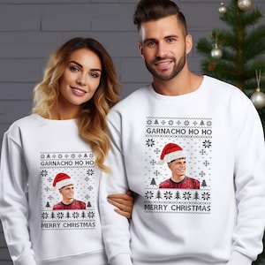 Könnte beinhalten: Weiße Sweatshirts mit festlichem Design. Jedes Sweatshirt zeigt eine Grafik einer Person mit Weihnachtsmütze, mit dem Text "GARNACHO HO HO" und "MERRY CHRISTMAS". Das Design umfasst Schneeflocken und Weihnachtsbäume.