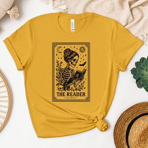 May include: Mustard yellow t-shirt with a black and white illustration of a skeleton reading a book, framed by floral elements, a crescent moon, and a sun. The text "THE READER" is below the illustration. The shirt is made of soft material.
