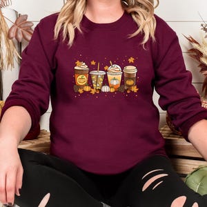 May include: Burgundy sweatshirt featuring a fall-themed graphic. The design includes four different coffee drinks with pumpkins, leaves, and fall colors. The sweatshirt is a long-sleeved crew neck.