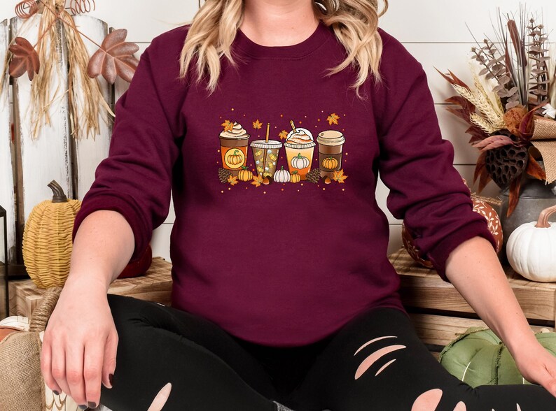 May include: Burgundy sweatshirt featuring a fall-themed graphic. The design includes four different coffee drinks with pumpkins, leaves, and fall colors. The sweatshirt is a long-sleeved crew neck.