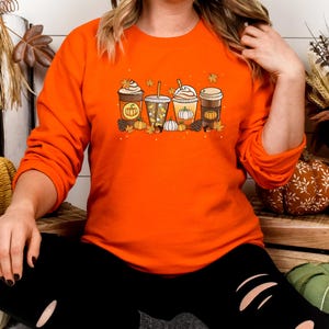 May include: Orange long-sleeved sweatshirt featuring a fall-themed graphic of four different coffee drinks with pumpkin and leaf accents. The design includes pumpkins and fall leaves. The sweatshirt is a solid orange color.
