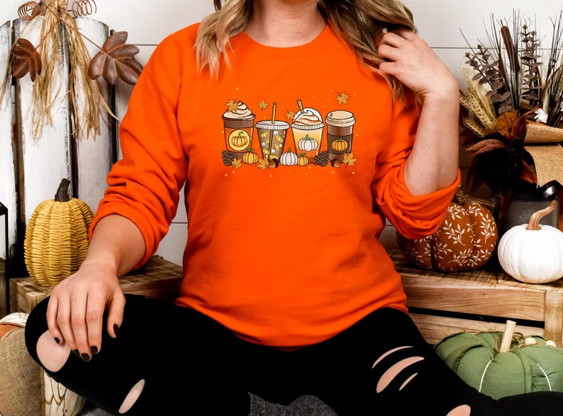 May include: Orange long-sleeved sweatshirt featuring a fall-themed graphic of four different coffee drinks with pumpkin and leaf accents. The design includes pumpkins and fall leaves. The sweatshirt is a solid orange color.
