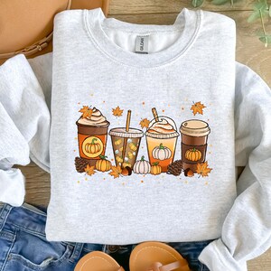 May include: Light gray sweatshirt featuring a colorful graphic of four fall-themed coffee drinks. Each drink has a pumpkin design, surrounded by autumn leaves, pumpkins, and pinecones. The sweatshirt is a crew neck style.