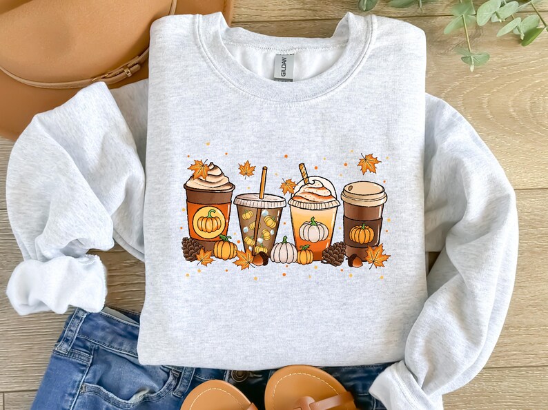 May include: Light gray sweatshirt featuring a colorful graphic of four fall-themed coffee drinks. Each drink has a pumpkin design, surrounded by autumn leaves, pumpkins, and pinecones. The sweatshirt is a crew neck style.