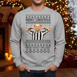 May include: A gray Christmas sweater with a festive design. The sweater features the text "MERRY CHRISTMAS" and "HO HO HOWAY THE LADS!" along with a graphic of a person in a black and white striped shirt.