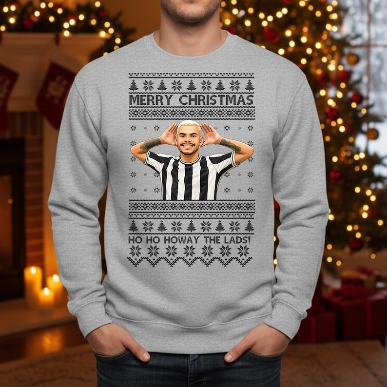 May include: A gray Christmas sweater with a festive design. The sweater features the text "MERRY CHRISTMAS" and "HO HO HOWAY THE LADS!" along with a graphic of a person in a black and white striped shirt.